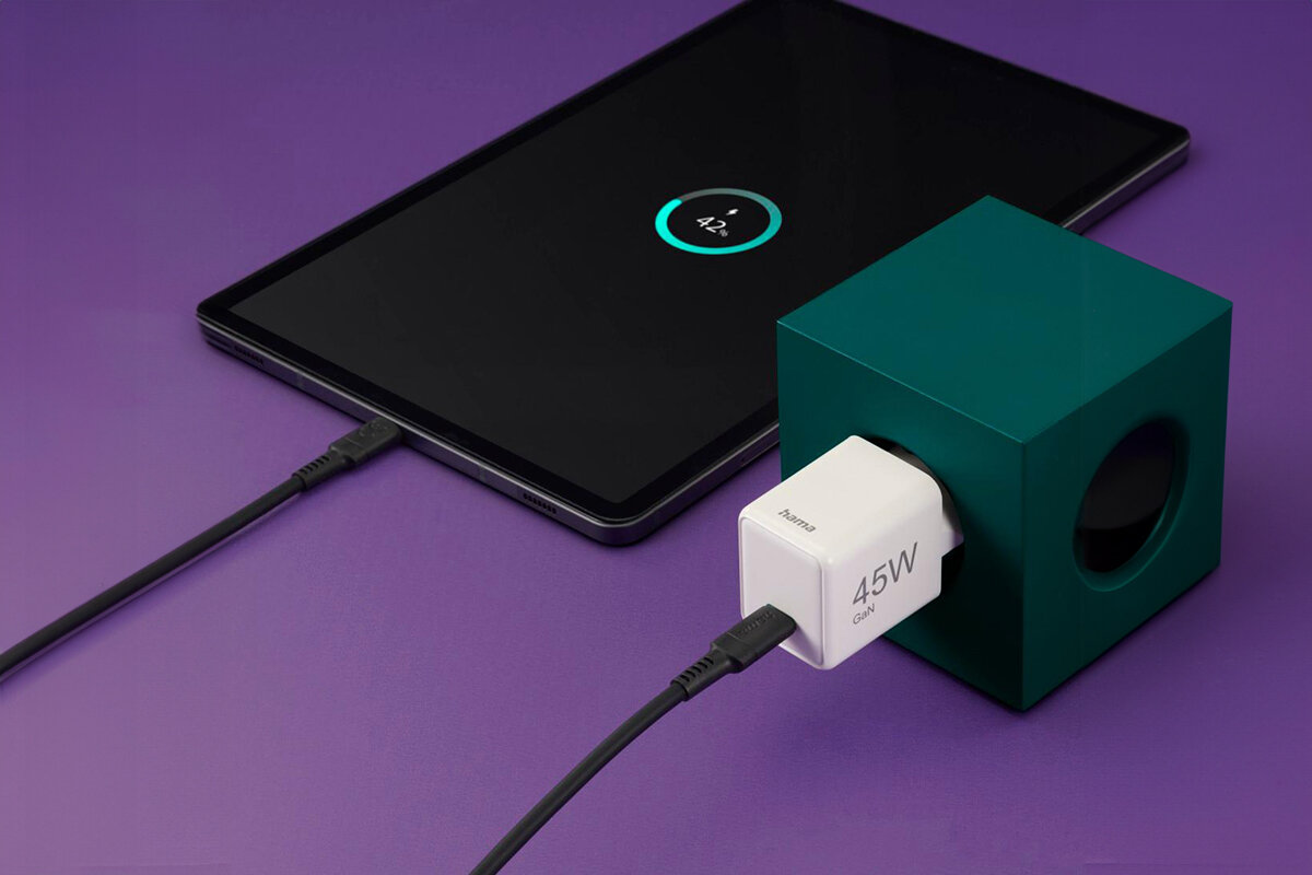 Na fioletowym tle znajduje się tablet z widocznym wskaźnikiem ładowania na ekranie oraz biała ładowarka z napisem \'Hama 45W GaN\' podłączona do zielonego sześcianu. Ładowarka jest połączona kablem USB-C z tabletem.