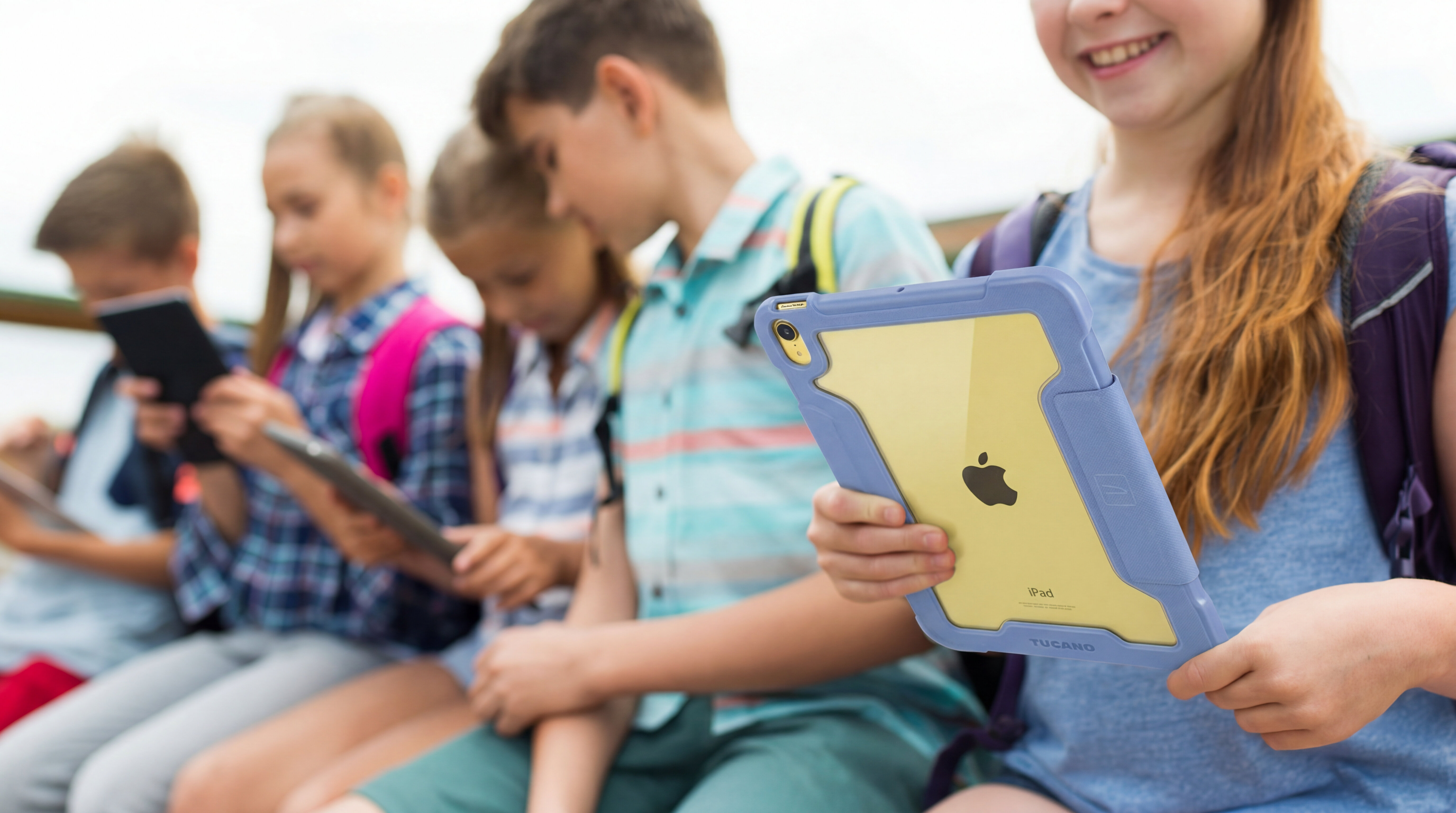 Grupa dzieci siedzi na ławce, każde z nich trzyma tablet w rękach. Jedno z dzieci trzyma tablet w niebieskim etui z widocznym logo Apple.
