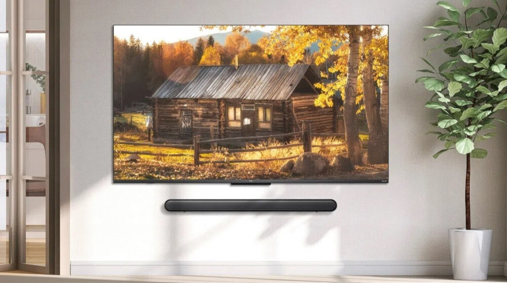 Na ścianie wisi duży telewizor wyświetlający obraz drewnianej chaty w jesiennym otoczeniu. Pod telewizorem znajduje się czarny soundbar, a obok stoi zielona roślina w doniczce.