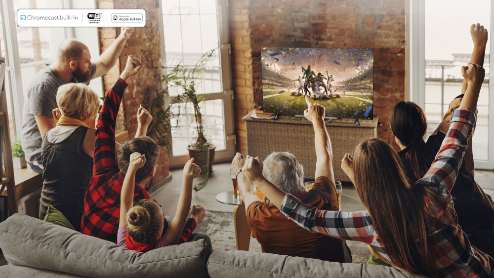 Grupa osób siedzi na kanapie w salonie, oglądając telewizor, na którym wyświetlany jest mecz piłkarski. W lewym górnym rogu widoczne są ikony: Chromecast built-in, WiFi, Apple AirPlay.