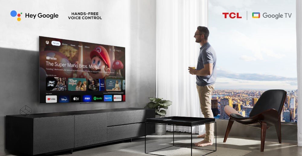 W salonie na ścianie wisi duży telewizor wyświetlający ekran startowy z aplikacjami, w tym YouTube z filmem \'The Super Mario Bros. Movie\'. Obok telewizora znajdują się logotypy \'Hey Google\', \'TCL\' i \'Google TV\'.