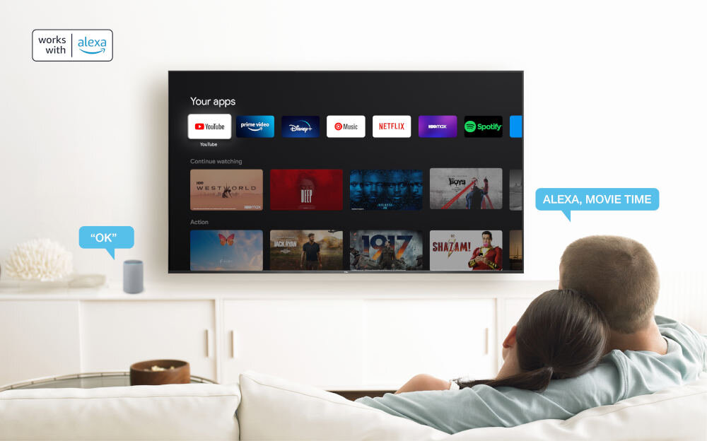 Na ścianie wisi duży telewizor z interfejsem aplikacji takich jak YouTube, Netflix i Spotify. Obok telewizora widoczny jest napis \'works with alexa\', a na ekranie napis \'ALEXA, MOVIE TIME\'.
