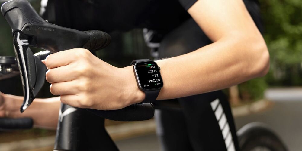 Osoba trzyma kierownicę roweru, na nadgarstku ma smartwatch z wyświetlaczem pokazującym czas i inne dane. Smartwatch ma czarny pasek i prostokątny ekran.