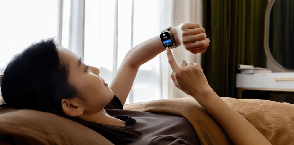 Osoba leży na łóżku i patrzy na smartwatch na swoim nadgarstku. Smartwatch wyświetla ekran z ikonami i tekstem.