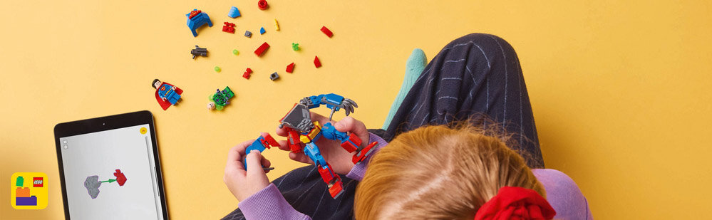 Dziecko siedzi na podłodze i trzyma w rękach złożony model LEGO, obok leży tablet z aplikacją pokazującą instrukcję składania. Na podłodze rozrzucone są różnokolorowe klocki LEGO.