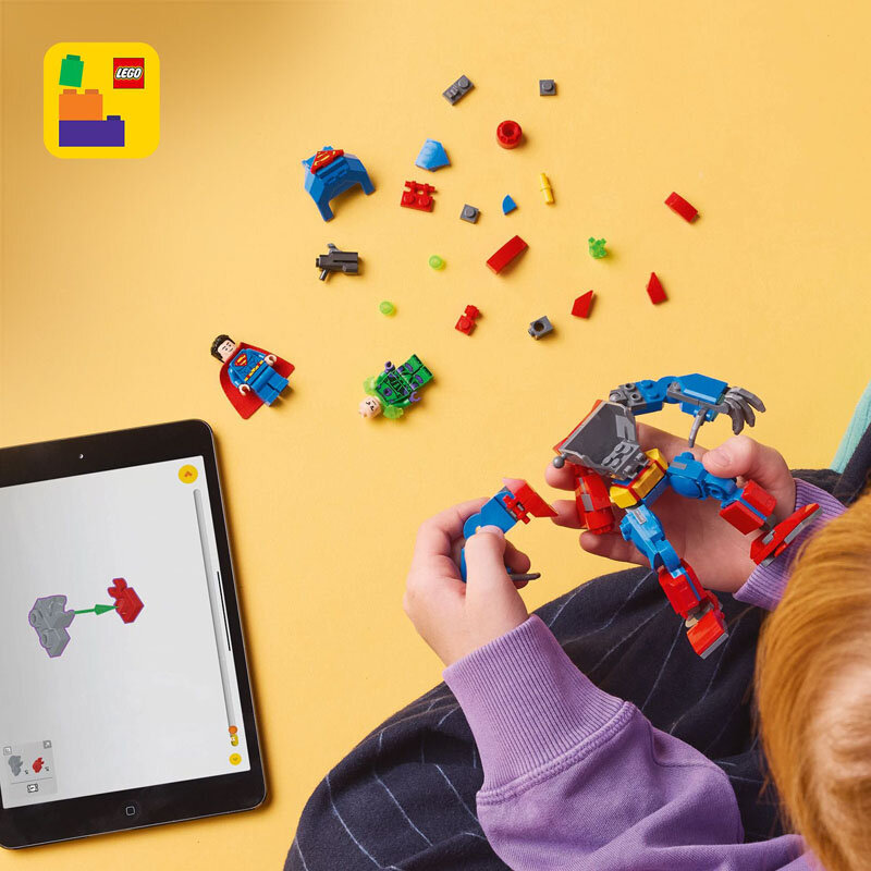 Na żółtym tle widoczne są elementy zestawu LEGO, w tym figurki i części konstrukcyjne. Obok znajduje się tablet z instrukcją budowy, a w rękach trzymana jest złożona część robota.