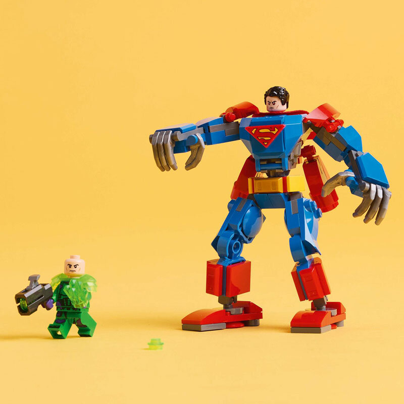 Na żółtym tle znajduje się duża figurka LEGO przedstawiająca mecha w kolorach niebieskim, czerwonym i szarym z symbolem \'S\' na piersi oraz mniejsza figurka w zielonym stroju z bronią. Obie figurki są wykonane z klocków LEGO.