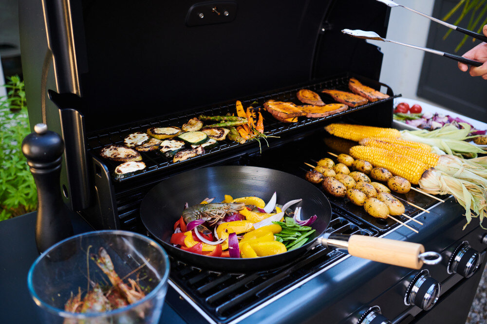 Na otwartym grillu gazowym znajdują się różnorodne warzywa, w tym cukinia, papryka, cebula, kukurydza i ziemniaki. Obok grilla stoi szklana miska z krewetkami oraz młynek do pieprzu.