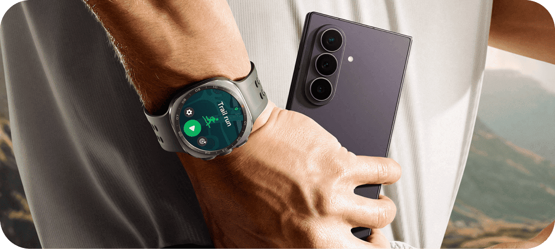 Galaxy Watch Ultra2 na nadgarstku i Galaxy Z Fold8 Ultra trzymany w dłoni. Osoba trzyma smartfon z trzema obiektywami aparatu, a na nadgarstku ma zegarek z wyświetlaczem pokazującym aplikację do biegania z tekstem \'Trail run\'.