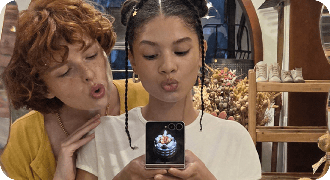 Dwie osoby robią selfie Galaxy Z Flip8, a na ekranie zewnętrznym widać tort. Dwie osoby robią sobie zdjęcie przy użyciu smartfona z widocznym ekranem, na którym wyświetla się obrazek tortu. W tle znajdują się półki z butami i dekoracje z suszonych kwiatów.