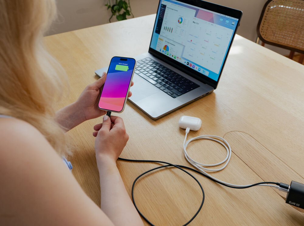 Osoba trzyma smartfon z kolorowym ekranem, podłączony do laptopa za pomocą kabla USB-C. Na ekranie laptopa widoczne są wykresy i dane, a obok leżą słuchawki bezprzewodowe.