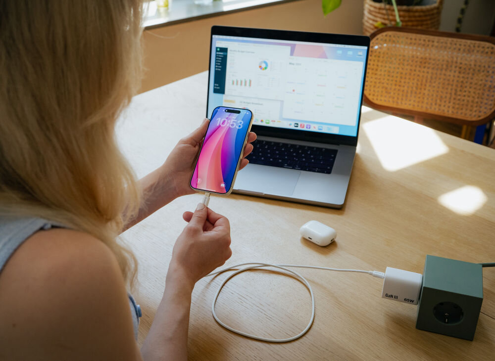 Osoba trzyma smartfon z wyświetlonym ekranem, podłączony do ładowania za pomocą kabla USB-C. W tle znajduje się otwarty laptop z widocznym interfejsem aplikacji oraz leżące na stole etui na słuchawki bezprzewodowe.