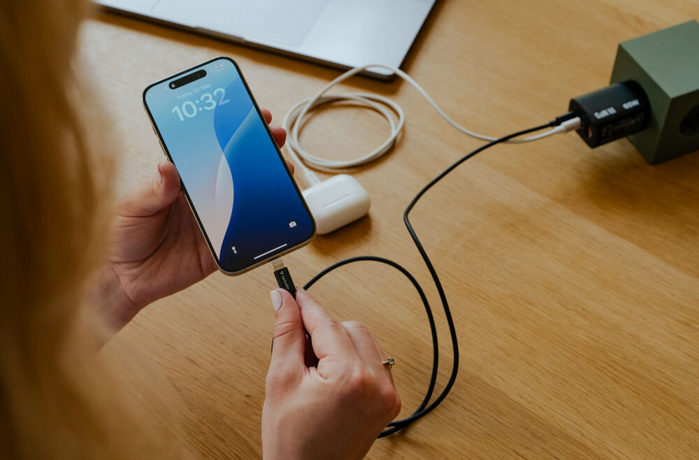 Osoba trzyma smartfon z wyświetlonym czasem 10:32, podłączając kabel USB-C do urządzenia. Na stole leży ładowarka i inne akcesoria elektroniczne.