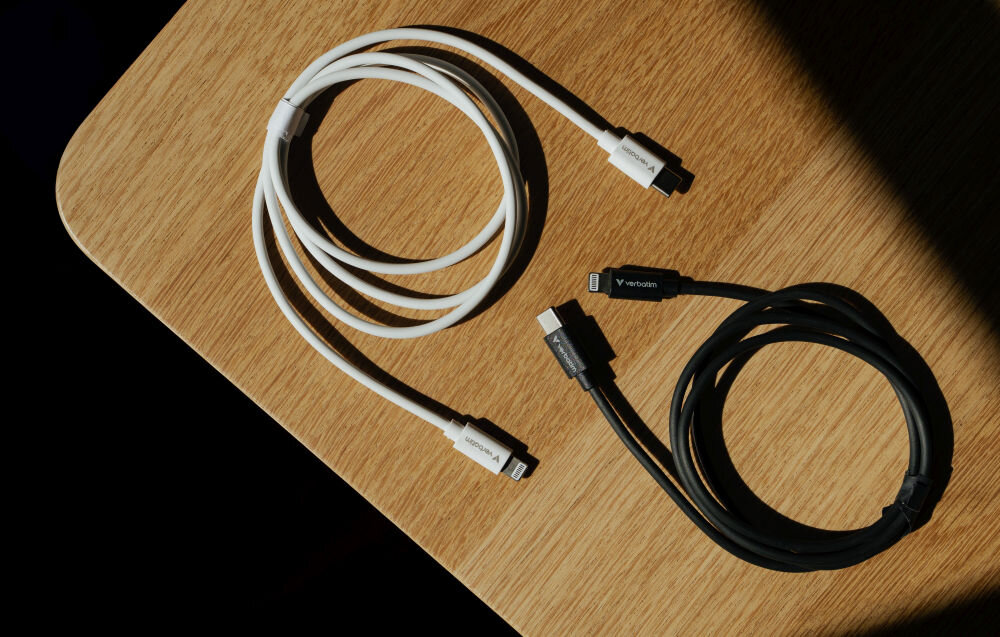 Na drewnianym blacie leżą dwa kable USB-C - Lightning, jeden biały i jeden czarny, oba z widocznym logo \'Verbatim\' na złączach.