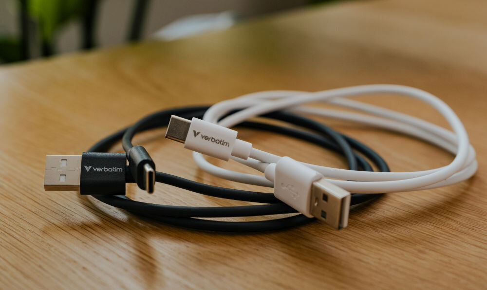 Na drewnianym stole leżą dwa kable USB-A do USB-C, jeden czarny i jeden biały, z widocznym logo \'verbatim\' na obu wtyczkach.