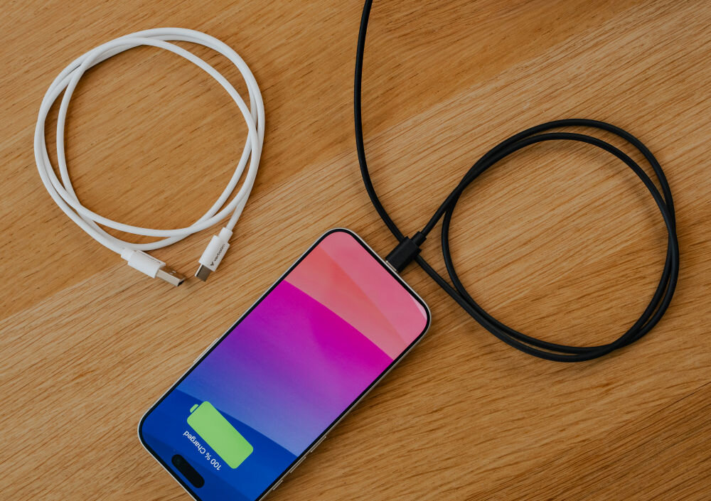 Na drewnianym blacie leży biały kabel USB-A do USB-C oraz czarny kabel podłączony do smartfona z kolorowym ekranem i ikoną ładowania baterii.
