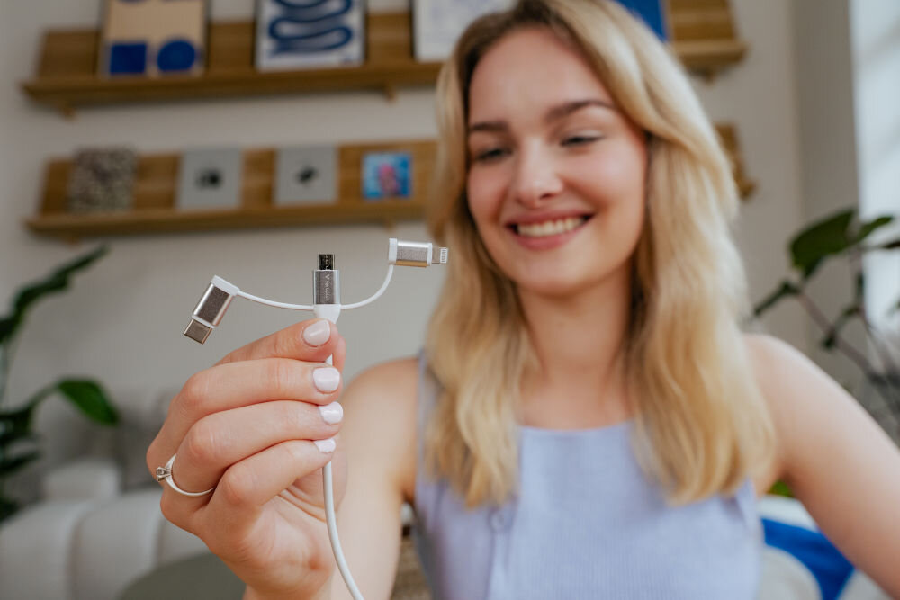 Osoba trzyma biały kabel z trzema różnymi końcówkami: USB-C, Micro USB i Lightning. W tle widoczne są półki z dekoracjami.