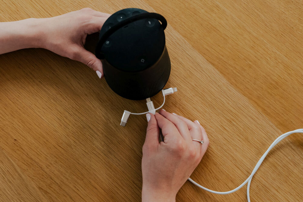 Osoba trzyma biały kabel z trzema różnymi końcówkami, obok znajduje się czarny głośnik na drewnianym stole.