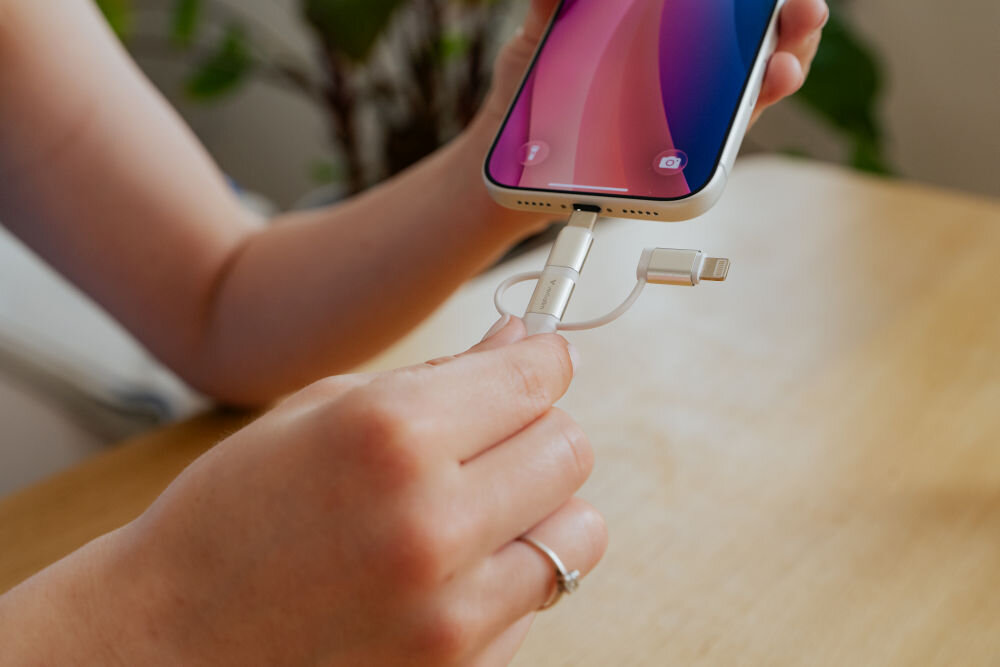 Osoba trzyma telefon i podłącza do niego biały kabel z wymiennymi końcówkami USB-C, Micro USB i Lightning. W tle widać rozmytą roślinę na drewnianym stole.
