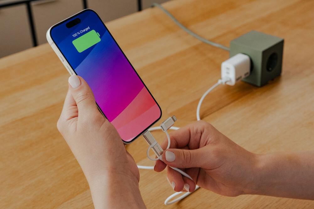 Osoba trzyma smartfon z wyświetlonym napisem \'100% Charged\' oraz kabel z trzema różnymi końcówkami. W tle widać ładowarkę podłączoną do gniazdka na drewnianym stole.