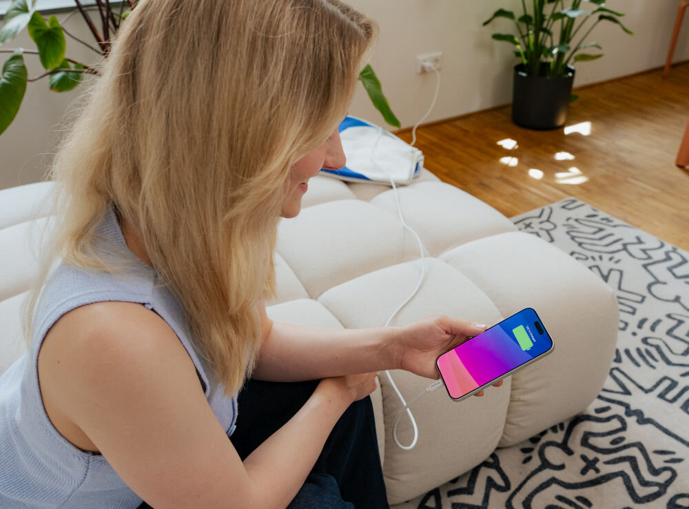 Osoba siedzi na jasnej kanapie, trzymając w ręku smartfon z kolorowym ekranem. Obok widoczny jest biały kabel podłączony do telefonu.