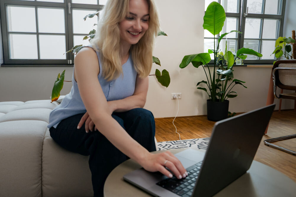 Osoba siedzi na kanapie i korzysta z laptopa, w tle widać rośliny doniczkowe i okno. Na podłodze leży dywan z geometrycznym wzorem.