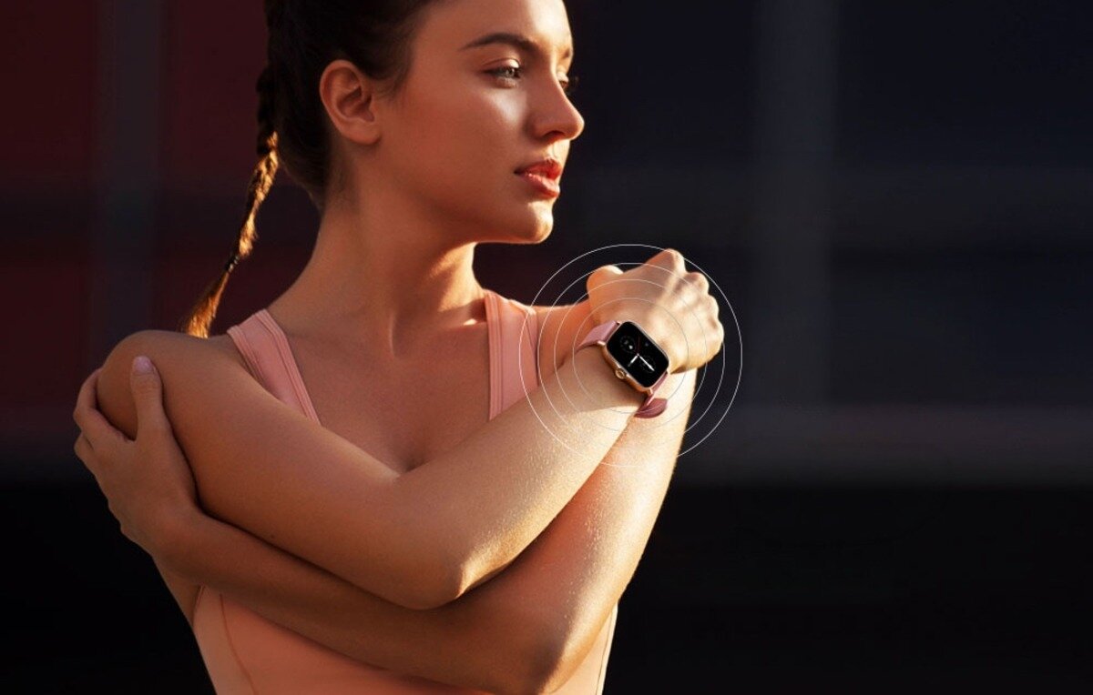 Osoba w sportowym stroju nosi srebrny smartwatch na nadgarstku. W tle widoczne jest rozmyte otoczenie.