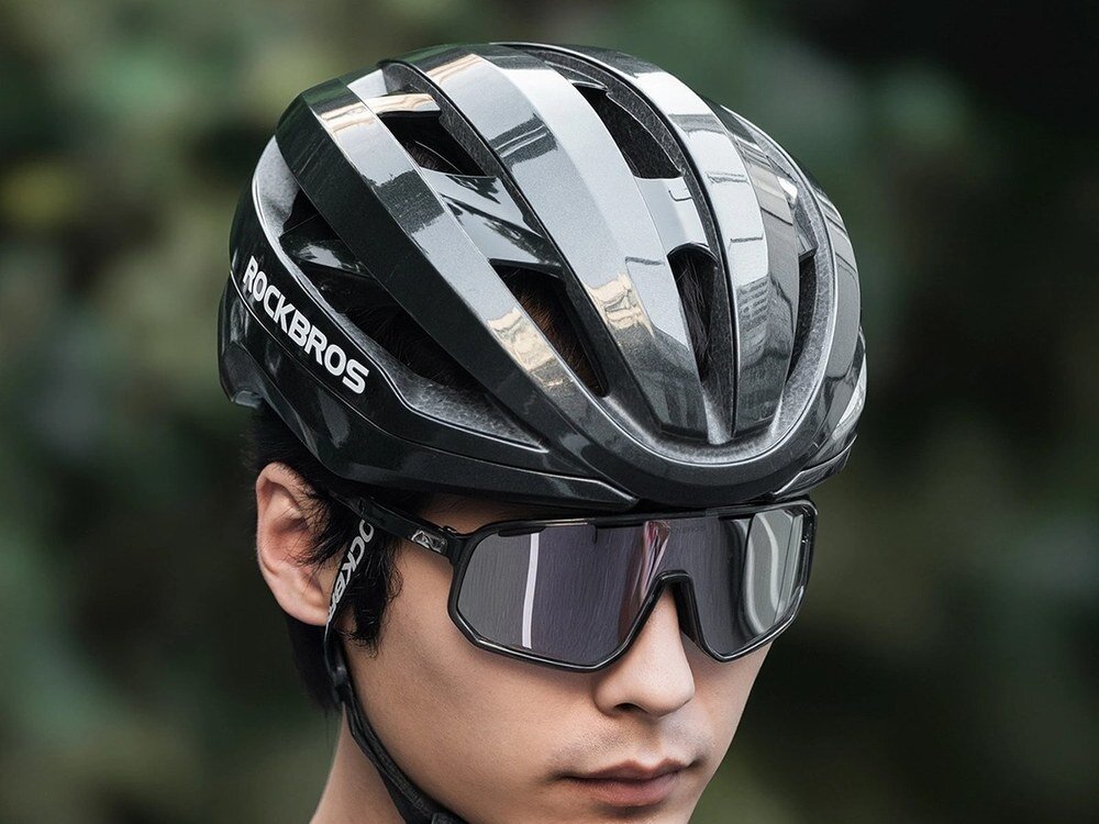 Osoba nosi czarny kask rowerowy z napisem \'ROCKBROS\' oraz okulary przeciwsłoneczne. Tło jest rozmyte, w zielonych odcieniach.