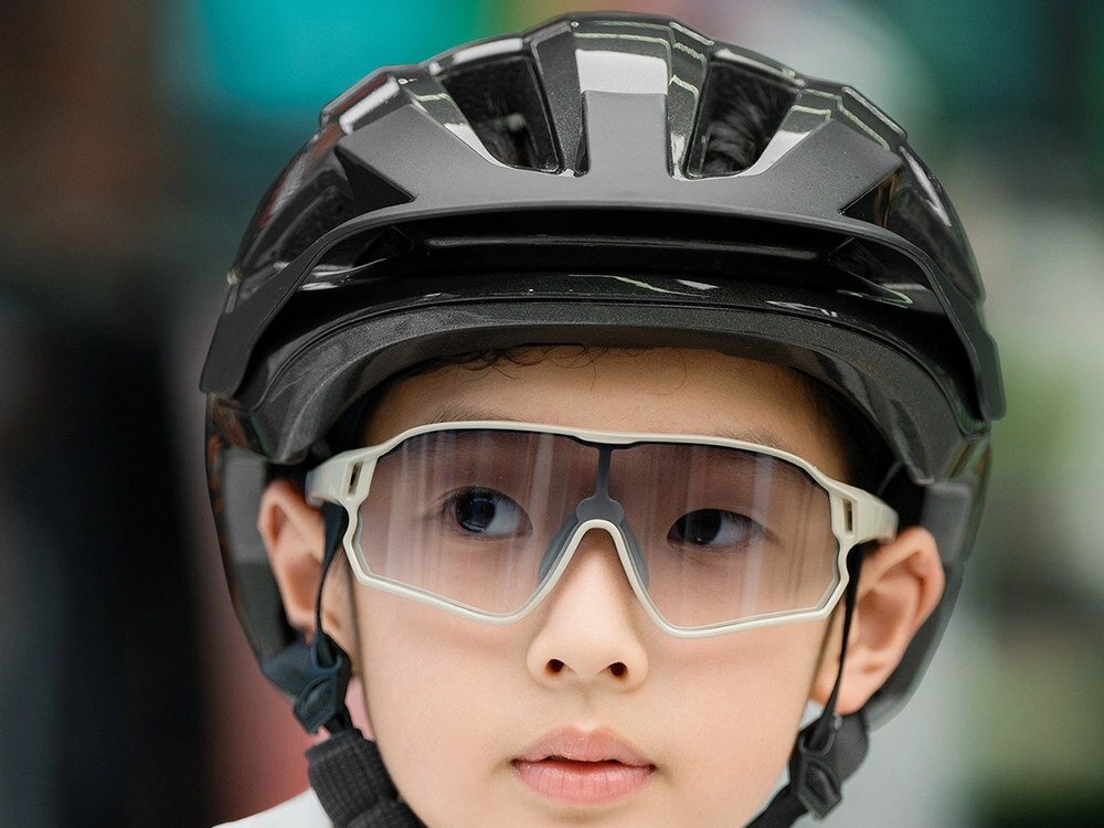 Osoba nosi czarny kask rowerowy i przezroczyste okulary ochronne. Tło jest rozmyte, co sugeruje, że zdjęcie skupia się na kasku.