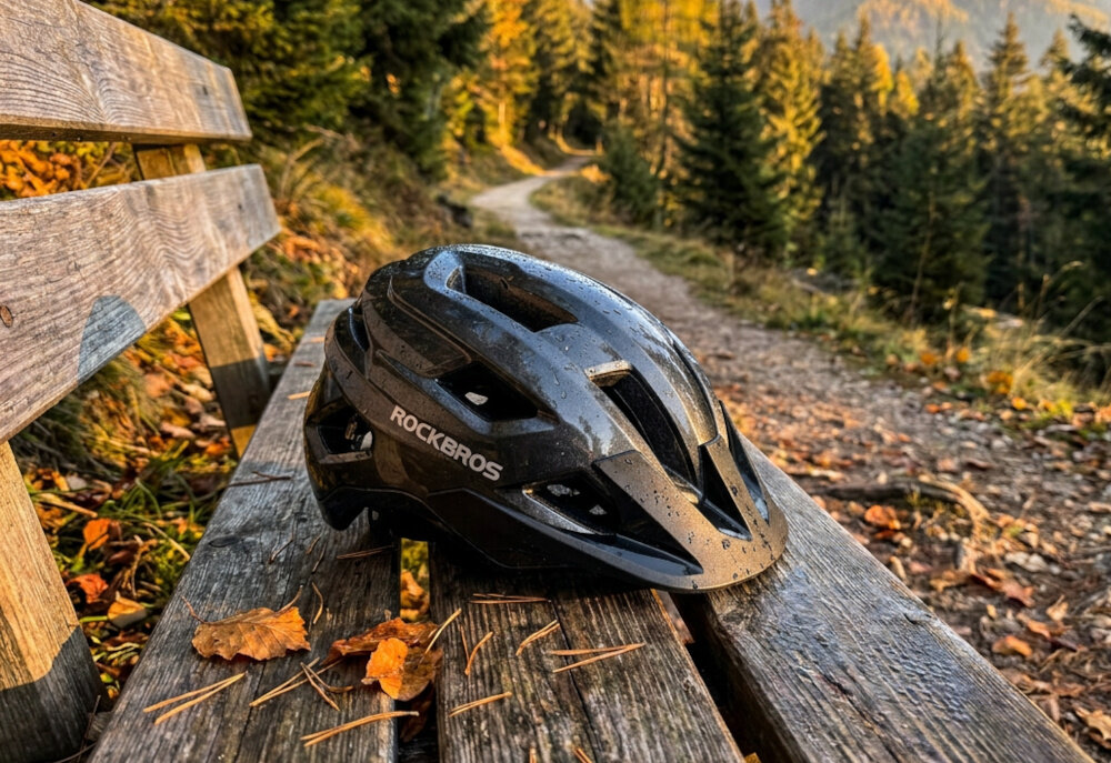 Czarny kask rowerowy z logo ROCKBROS leży na drewnianej ławce w otoczeniu jesiennych liści. W tle widoczna jest leśna ścieżka i drzewa.
