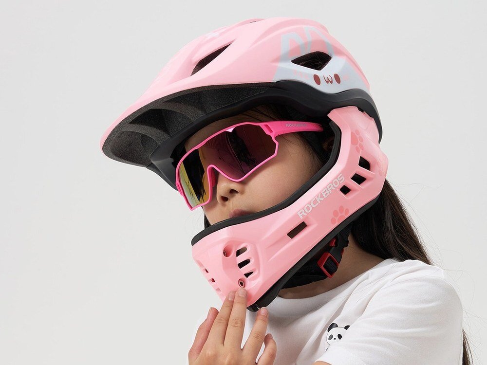 Dziecko nosi różowy kask rowerowy z odkręcaną szczęką i różowe okulary przeciwsłoneczne. Na kasku widoczne jest logo \'ROCKBROS\'.