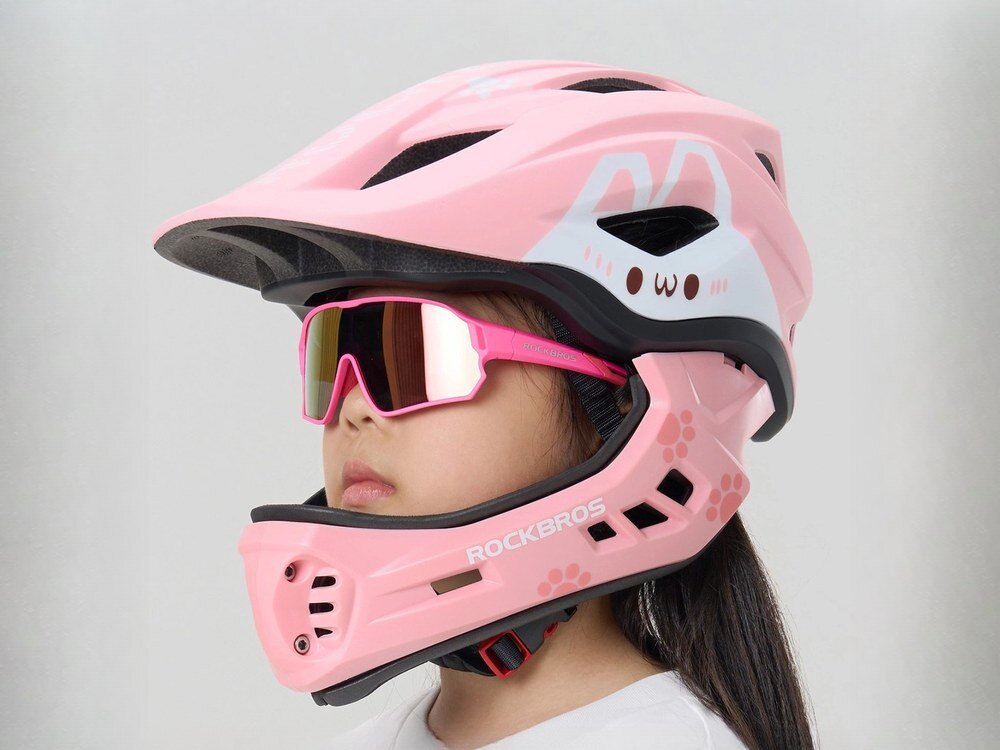 Dziecko nosi różowy kask rowerowy z odkręcaną szczęką, ozdobiony motywem króliczka i napisem \'ROCKBROS\'. Kask jest połączony z różowymi okularami przeciwsłonecznymi.