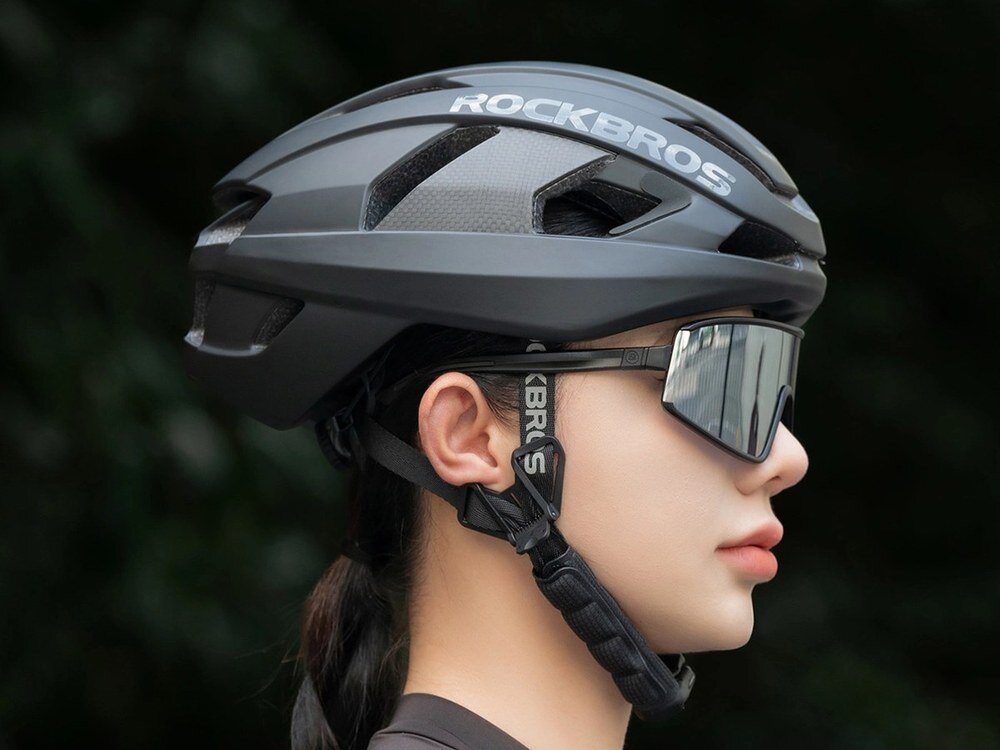 Ciemnoszary kask rowerowy Rockbros LK-15 na głowie kobiety widziany z boku, z widocznymi otworami wentylacyjnymi i zapiętymi paskami, w tle zieleń.
