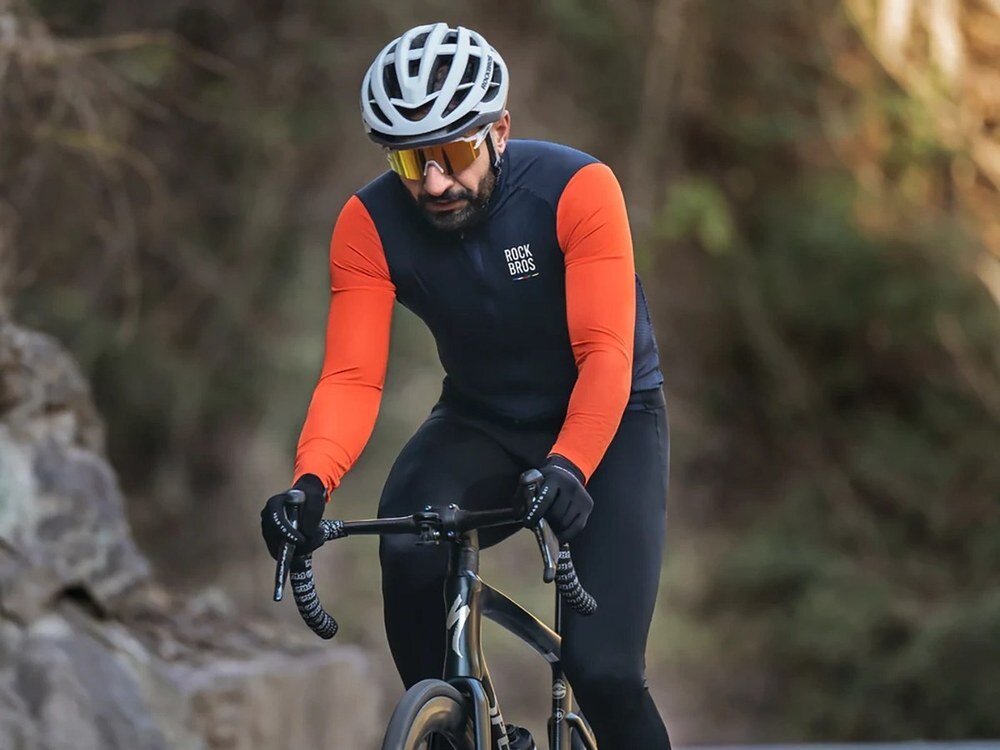 Mężczyzna w białym kasku Rockbros i okularach sportowych jedzie na rowerze szosowym, ubrany w granatowo-pomarańczową bluzę, w tle skały i zieleń.