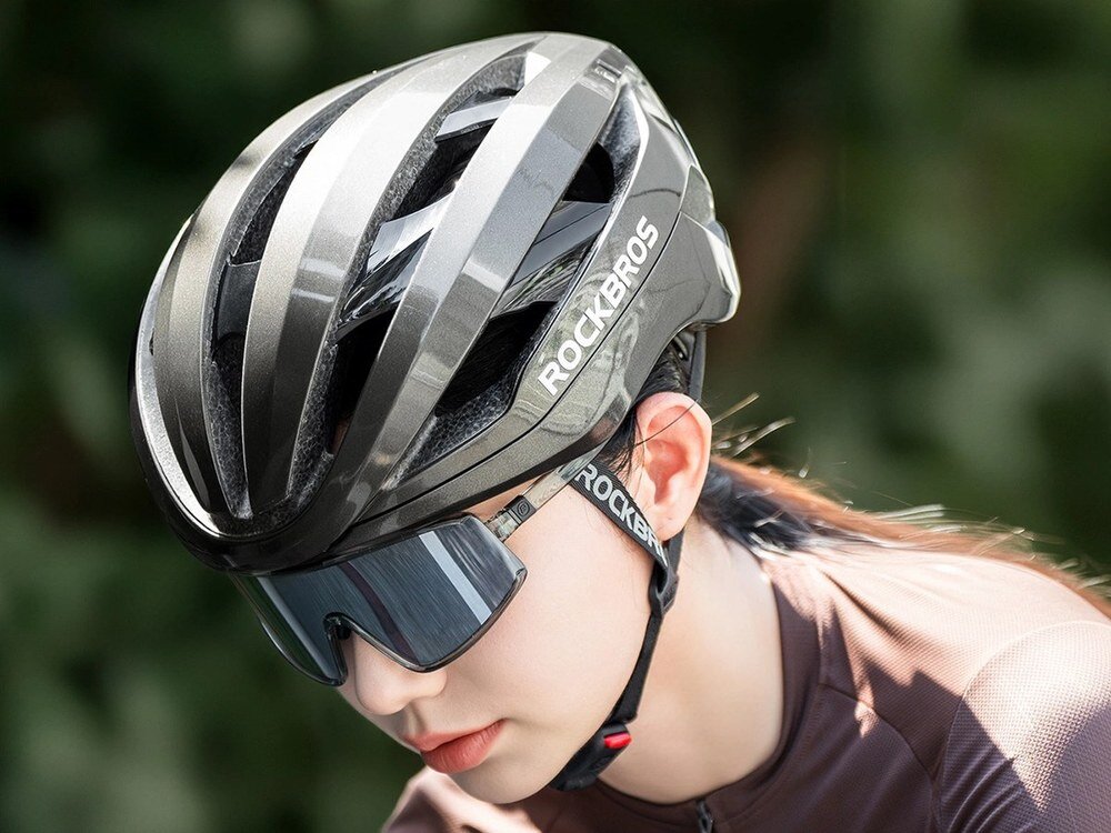 Osoba nosi szary kask rowerowy z napisem \'ROCKBROS\' oraz okulary przeciwsłoneczne. W tle widoczne są rozmyte zielone liście.