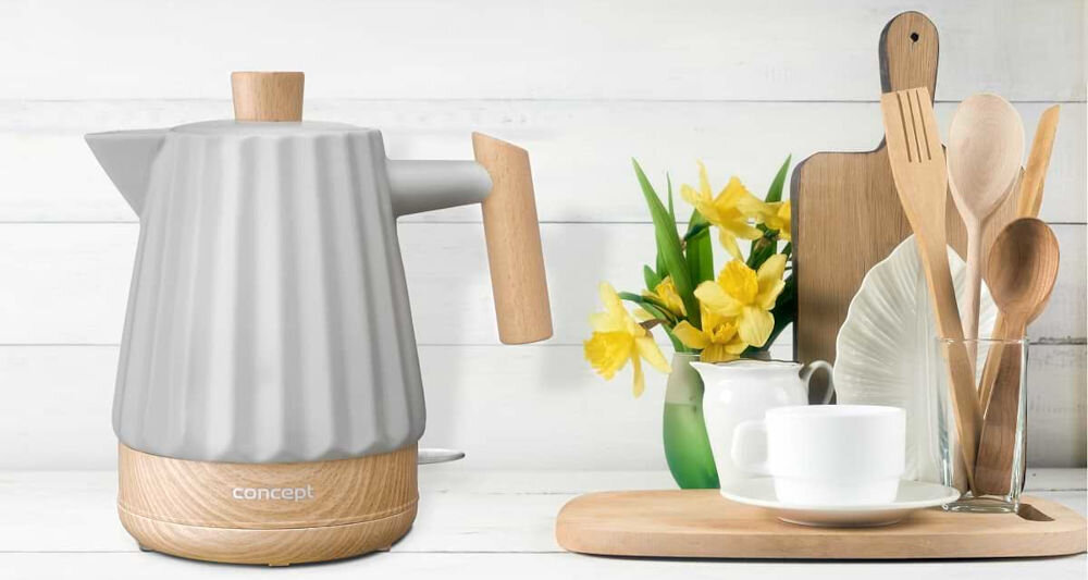 Na obrazie widoczny jest jasnoszary czajnik ceramiczny z drewnianymi elementami, obok którego znajduje się zestaw drewnianych przyborów kuchennych i filiżanka na drewnianej desce. Na czajniku widnieje napis \'concept\'.