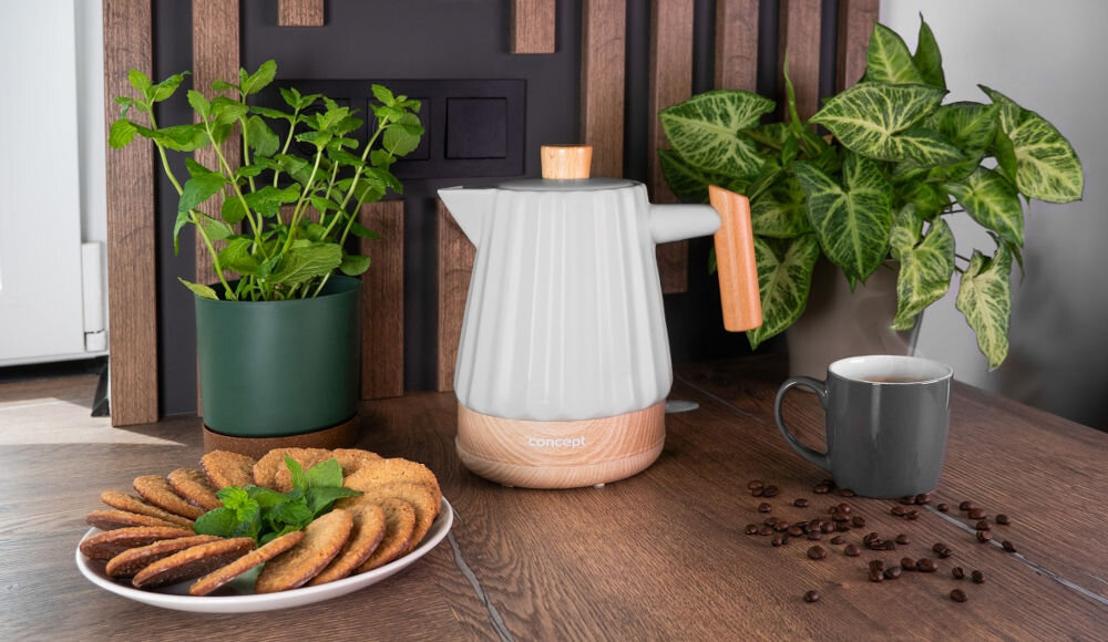 Na drewnianym blacie stoi jasnoszary czajnik ceramiczny z drewnianymi elementami, obok którego znajduje się filiżanka z kawą i talerz z ciastkami. W tle widoczne są zielone rośliny doniczkowe oraz kilka ziaren kawy rozsypanych na blacie.
