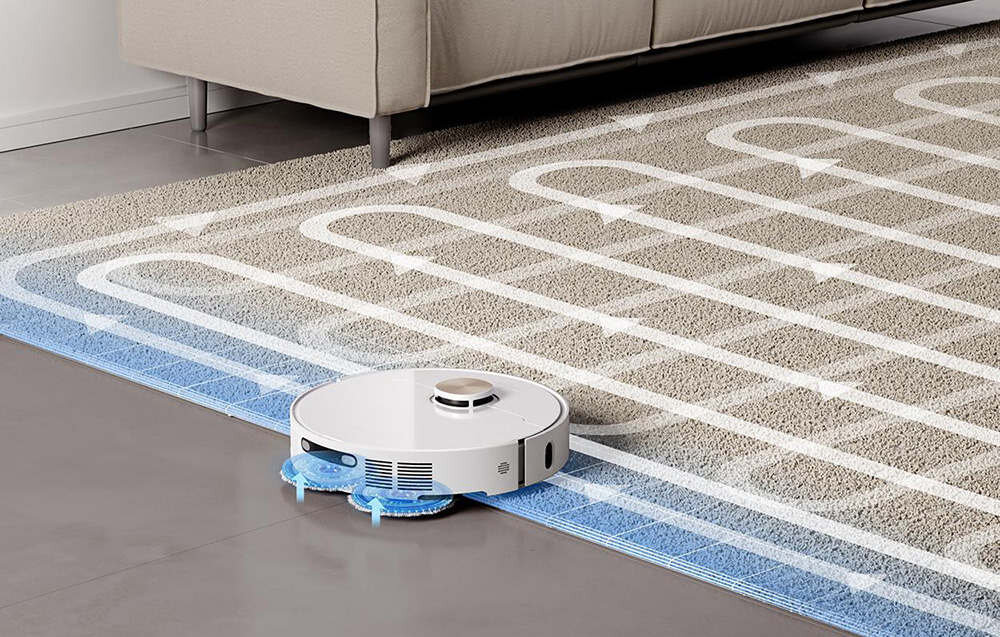 Na obrazie widoczny jest biały robot sprzątający na dywanie z wzorem w białe linie i strzałki. W tle znajduje się fragment kanapy na podłodze z szarych płytek.