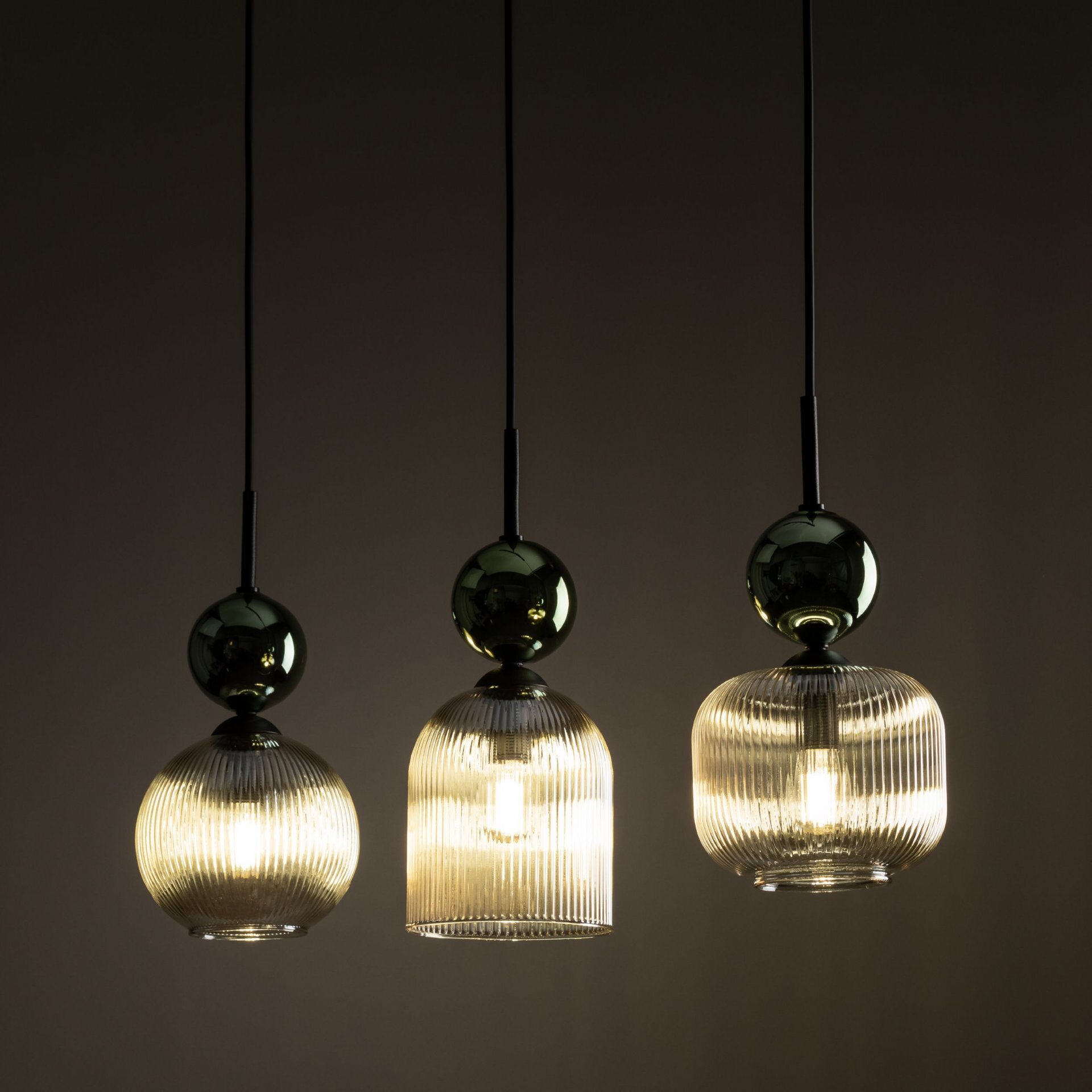 Trzy wiszące lampy z przezroczystymi, żebrowanymi kloszami, każda z metalicznym elementem na górze. Lampy emitują ciepłe, rozproszone światło.