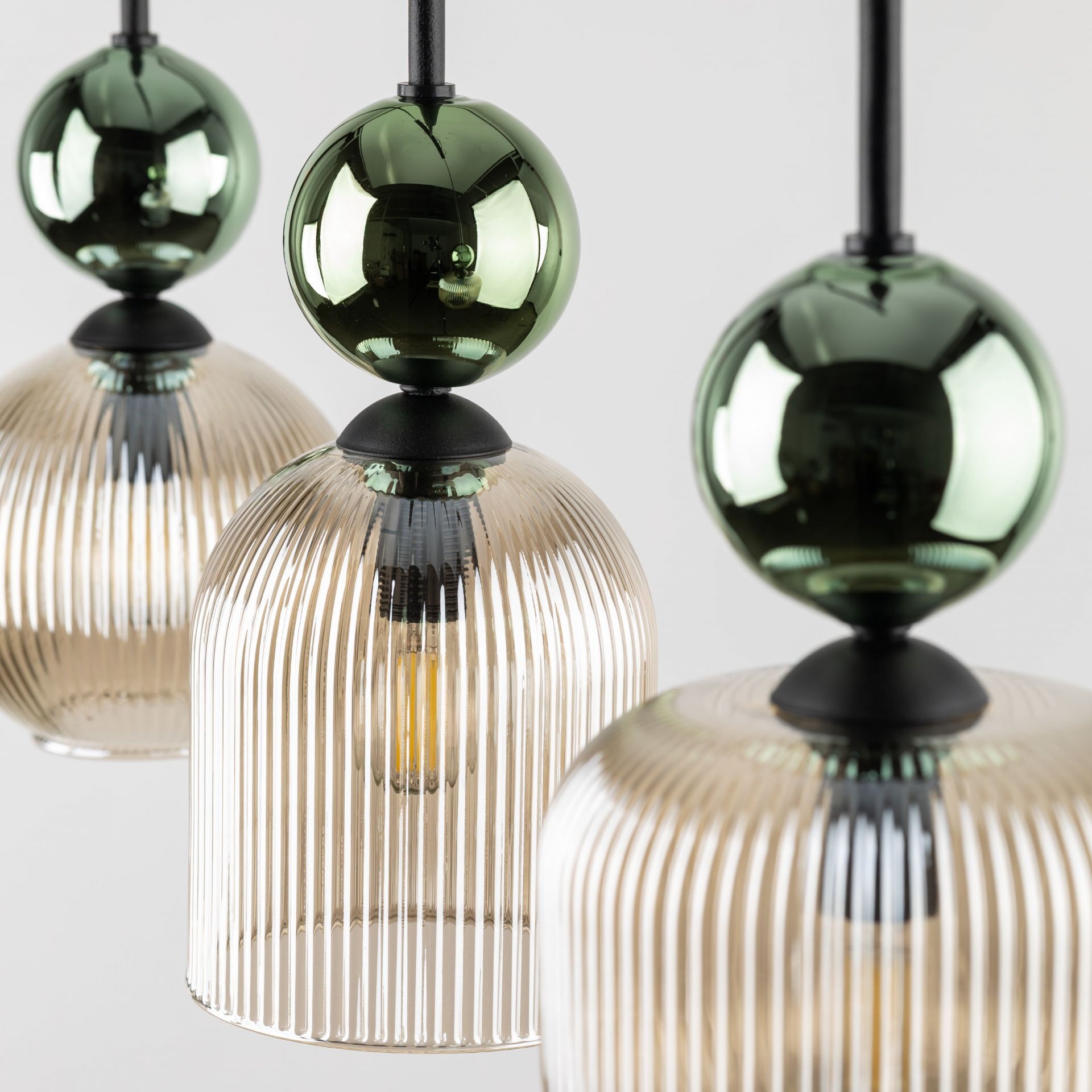 Zdjęcie przedstawia trzy wiszące lampy z przezroczystymi, żebrowanymi kloszami w kolorze koniakowym i zielonymi, błyszczącymi kulami na górze. Lampy są zamontowane na czarnych przewodach.