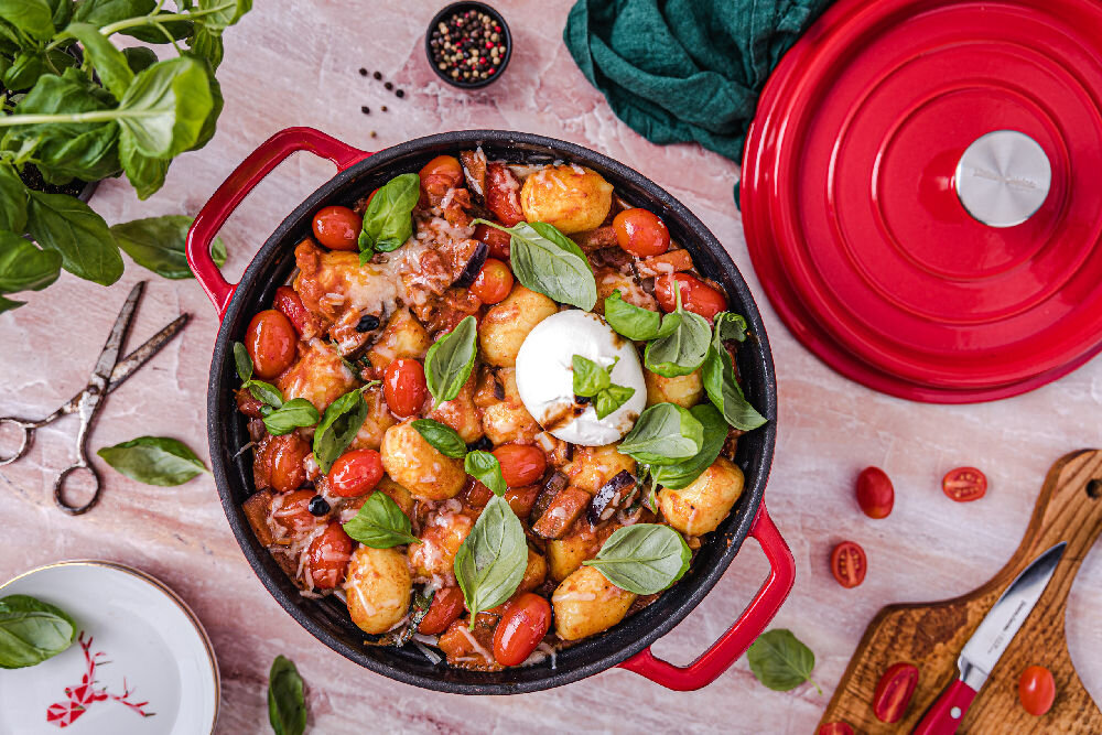 Na obrazie widoczny jest żeliwny garnek z czerwonymi uchwytami, wypełniony daniem z gnocchi, pomidorkami koktajlowymi, liśćmi bazylii i serem. Obok garnka leży czerwona pokrywka, zielona ściereczka oraz deska do krojenia z nożem i pomidorkami.