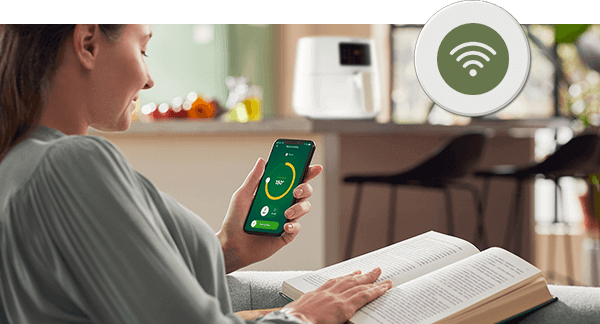 Osoba siedzi z książką, trzymając smartfon z aplikacją pokazującą okrągły wskaźnik. W tle na blacie kuchennym stoi biała frytkownica beztłuszczowa.