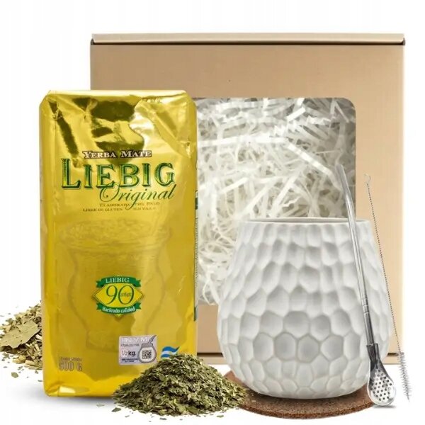 Na obrazie znajduje się złote opakowanie Yerba Mate Liebig Original, biały kubek o fakturze plastra miodu oraz metalowa bombilla. W tle widoczna jest kartonowa skrzynka wypełniona białym wypełniaczem.