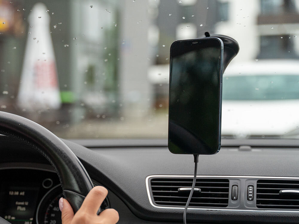 Wnętrze samochodu z zamontowanym na szybie uchwytem na telefon, w którym znajduje się smartfon. Widoczna jest kierownica oraz fragment deski rozdzielczej z kratką nawiewu.