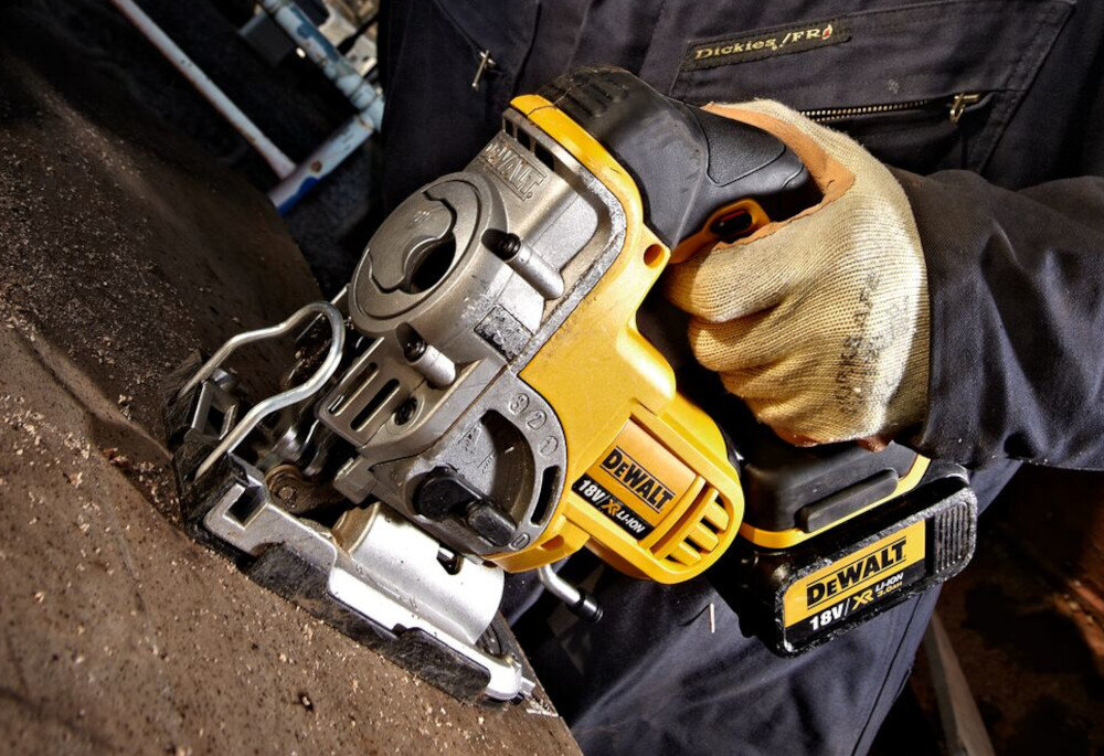 Osoba trzyma żółto-czarną wyrzynarkę marki DEWALT z widocznym logo i oznaczeniem \'18V XR Li-Ion\'. Narzędzie jest używane do cięcia powierzchni drewnianej.