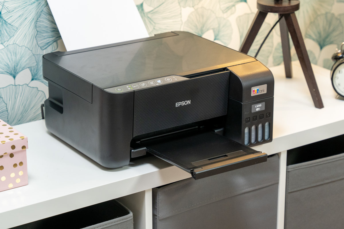 Na białej półce stoi czarne urządzenie wielofunkcyjne z logo EPSON, z wysuniętą tacką na papier. Obok urządzenia znajduje się pudełko w kropki i zegar, a w tle widoczna jest tapeta z motywem roślinnym.