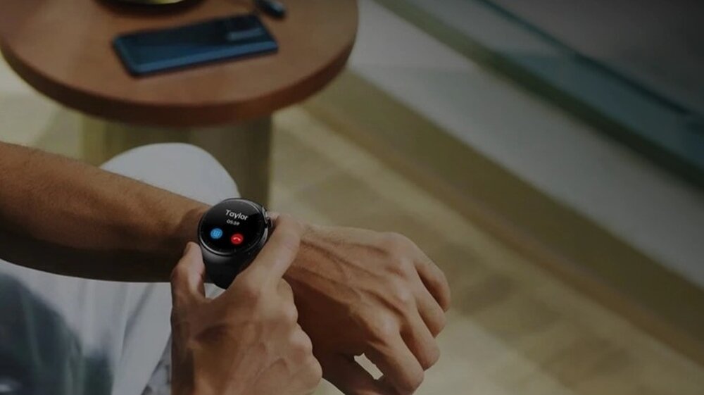 Osoba nosi smartwatch, na którego ekranie widoczny jest przychodzący telefon od \'Taylor\'. W tle znajduje się stół z telefonem komórkowym.
