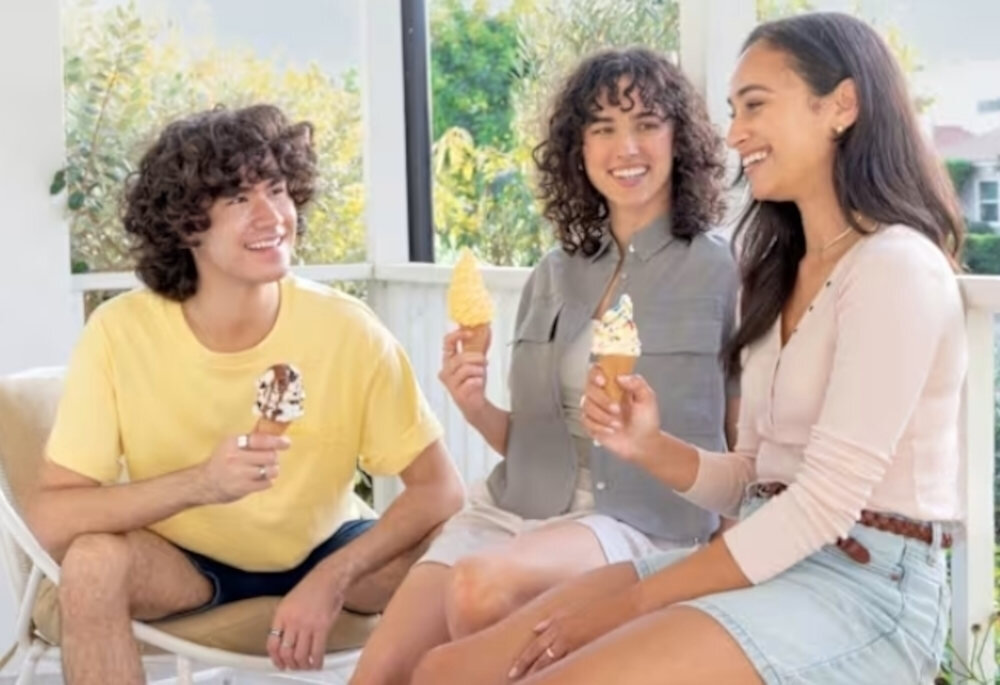 Trzy osoby siedzą na zewnątrz, każda trzyma lody w wafelku. W tle widać roślinność i fragment budynku.
