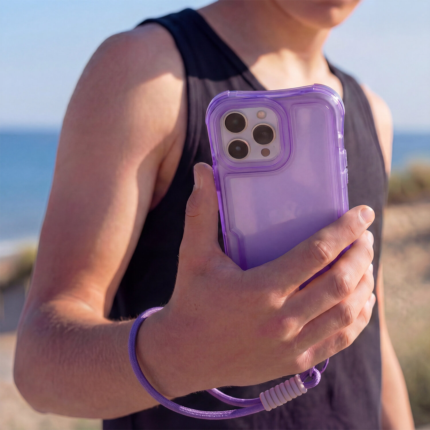 Osoba trzyma telefon w fioletowej nakładce z dołączoną smyczą. W tle widać plażę i morze.