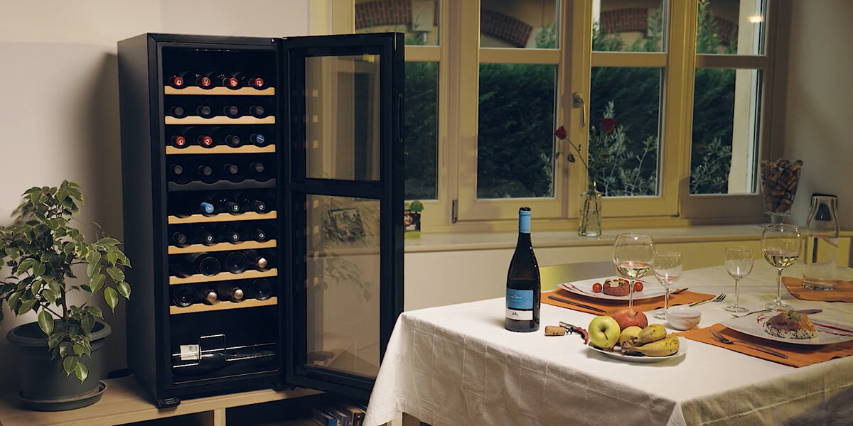 W jadalni stoi otwarta chłodziarka do wina Vermentino 36 C Dual z butelkami ułożonymi na drewnianych półkach. Obok znajduje się roślina oraz nakryty stół z winem i owocami 36 butelek 105 l dwie strefy 5-18°C